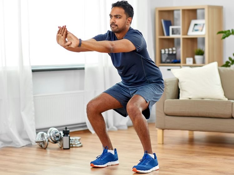 A beginner practicing a bodyweight squat during a home workout routine in a bright living room
